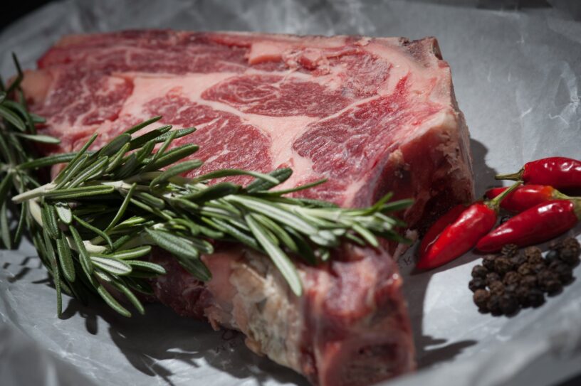 Raw steak, shows the different fibers meat is composed of (muscles, fats, blood vessels). Some herbs and peppers on the side for decoartion.